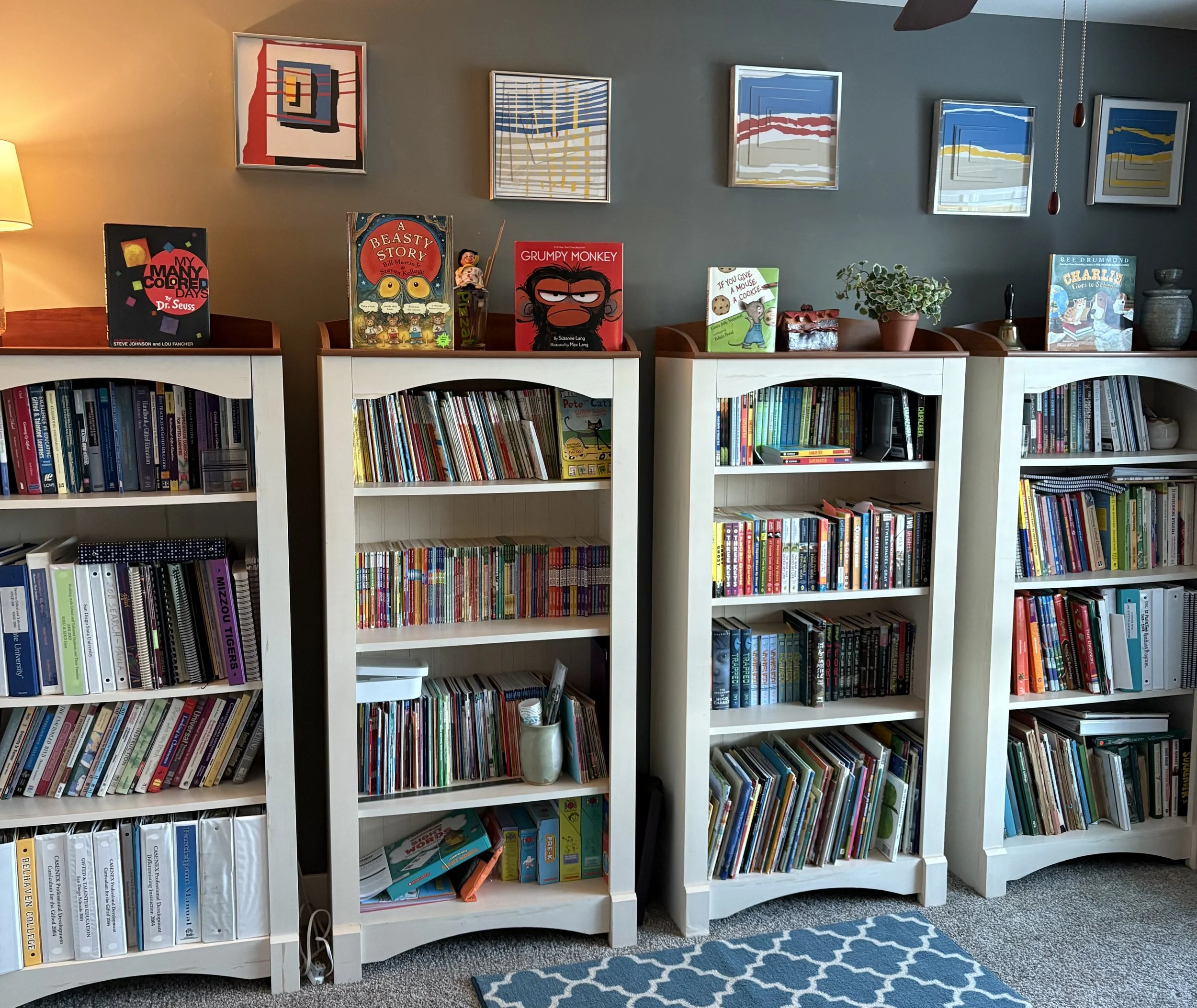 Bookshelves filled with children's books in a room with a patterned rug, artwork on the wall, and a beige carpet.