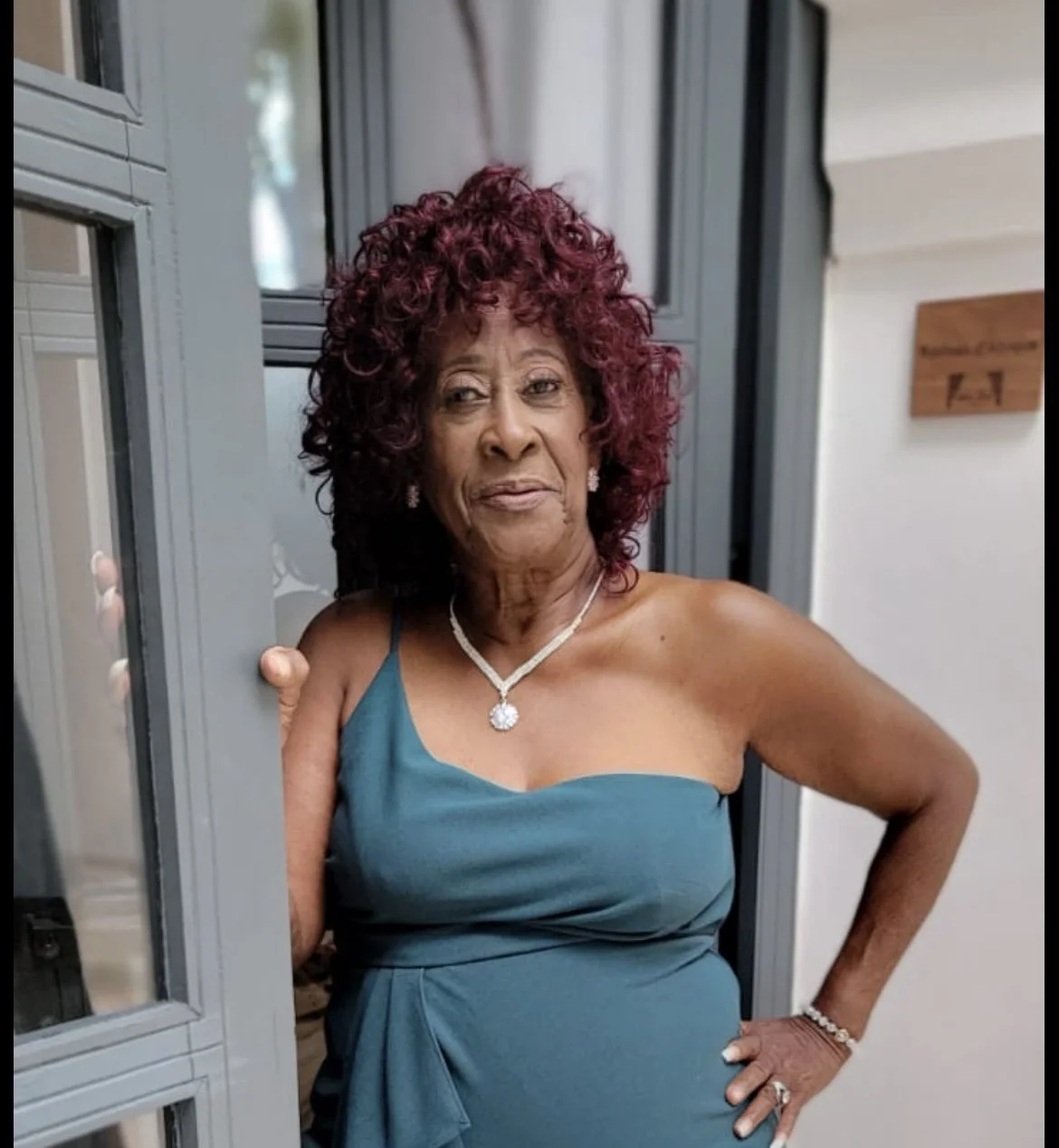 A woman with curly red hair wearing a teal dress, a necklace, earrings, and a bracelet, standing outside a building with her hand on her hip, smiling slightly.