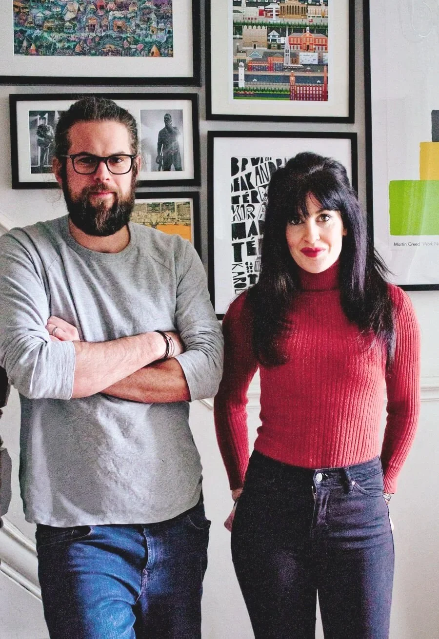 A man with glasses and a beard wearing a grey long-sleeve shirt, standing with arms crossed, next to a woman with black hair in a red ribbed turtleneck and black jeans, in front of a wall decorated with framed artwork.