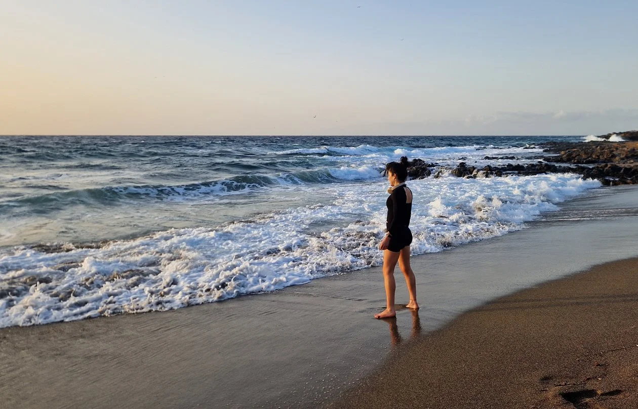 A woman standing barefoot on the beach at sunset, looking at the ocean waves.