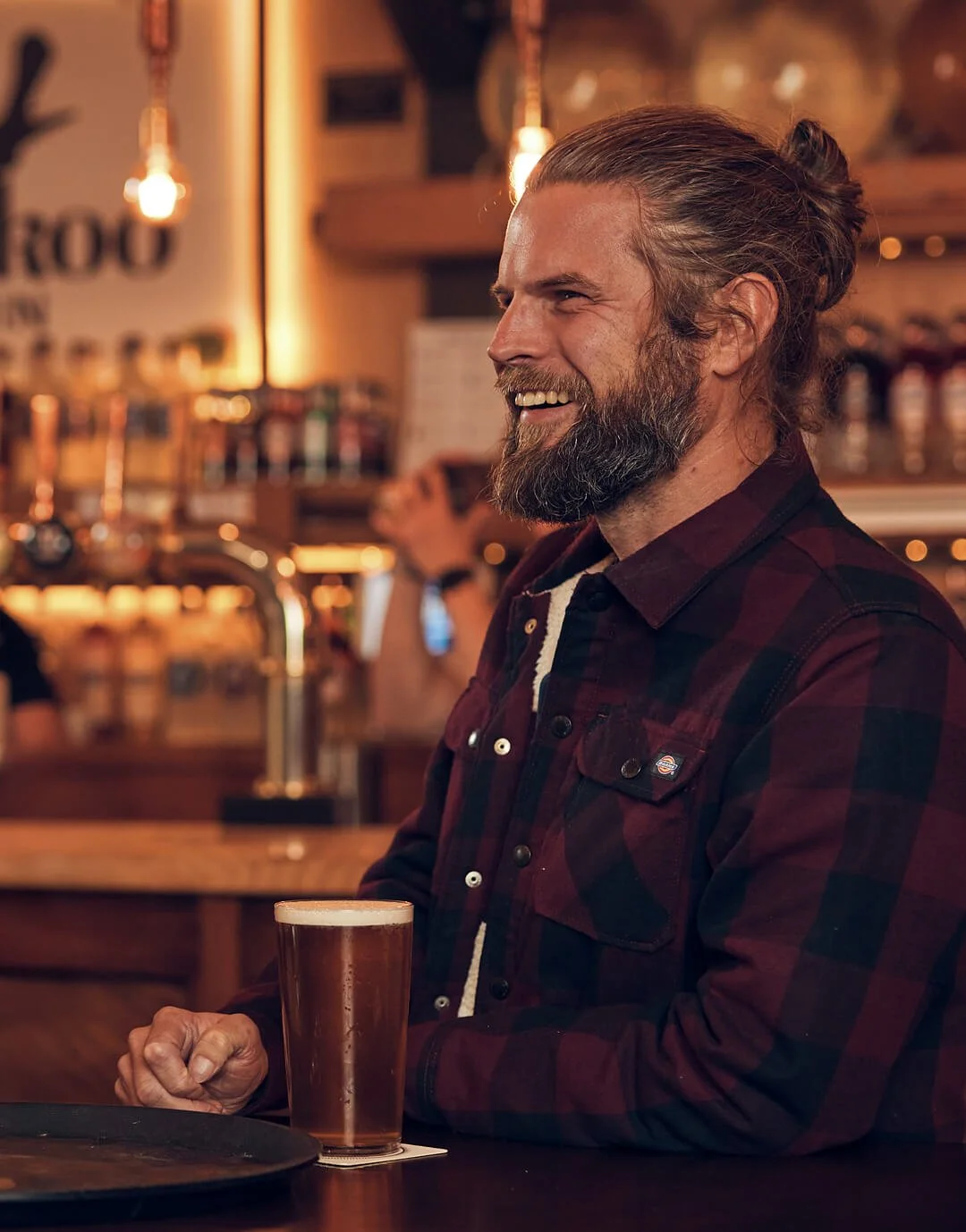 A man with a beard and long hair tied in a bun, wearing a red and black plaid shirt, sitting in a bar with a glass of beer in front of him, smiling.