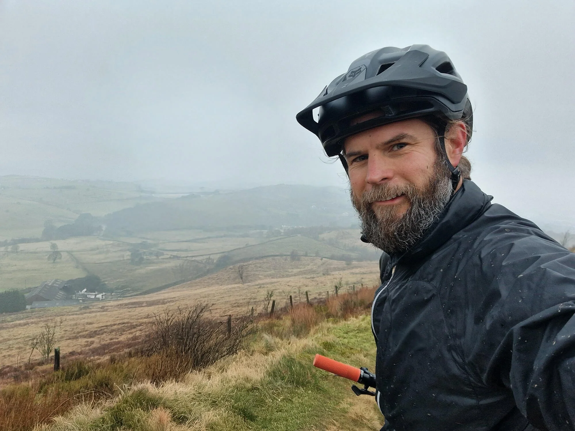 A man with a beard wearing a black bicycle helmet and black jacket taking a selfie outdoors on a cloudy day, with rolling hills and a fenced grassy field in the background.