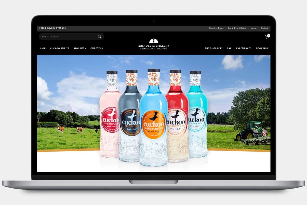Laptop screen displaying a website for Brindle Distillery with bottles of Cuckoo craft gin in front of an outdoor farm scene background.