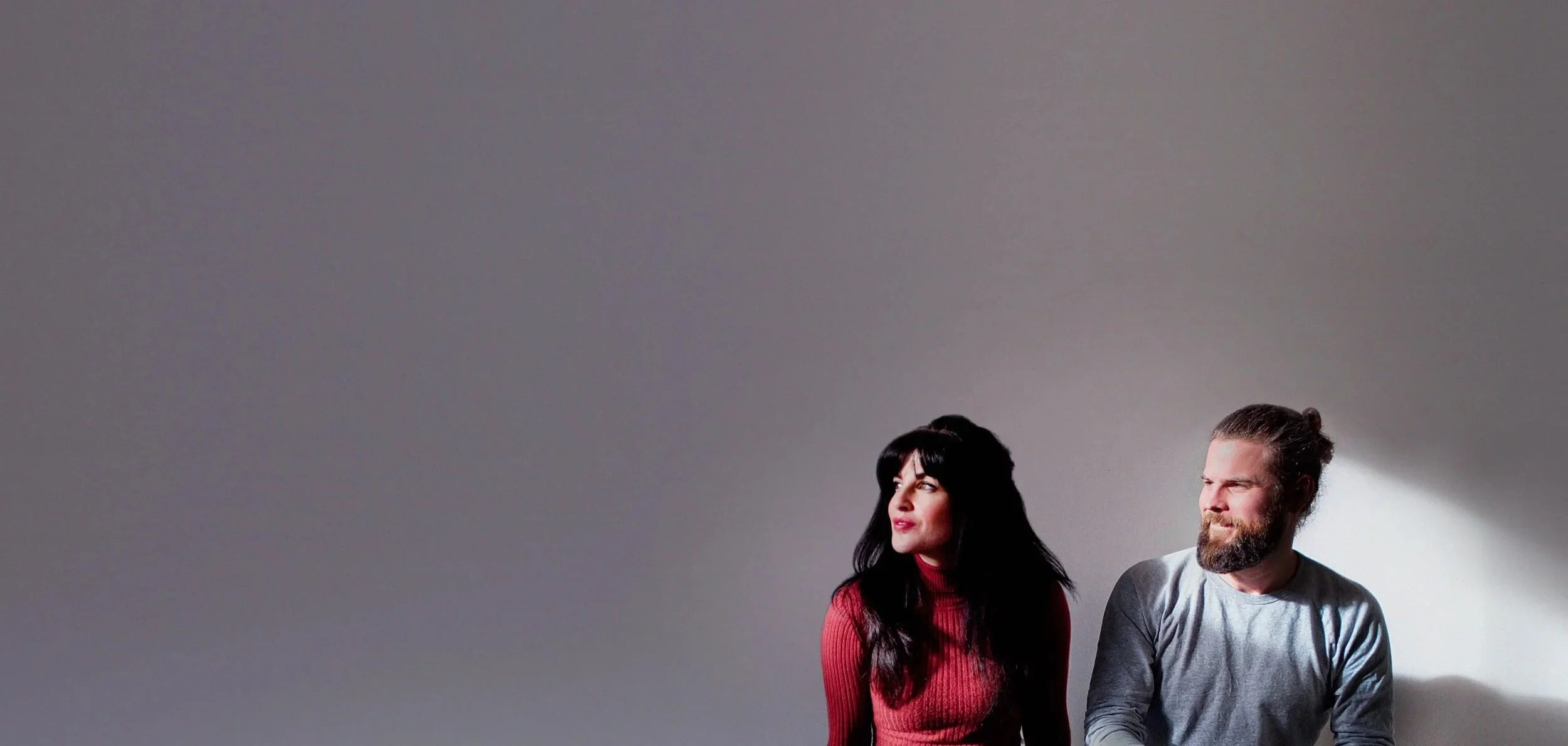 A woman and a man sitting against a plain grey background, looking slightly to their left with neutral expressions. The woman has long dark hair and is wearing a red top, while the man has a beard and long hair tied back, wearing a light grey shirt.