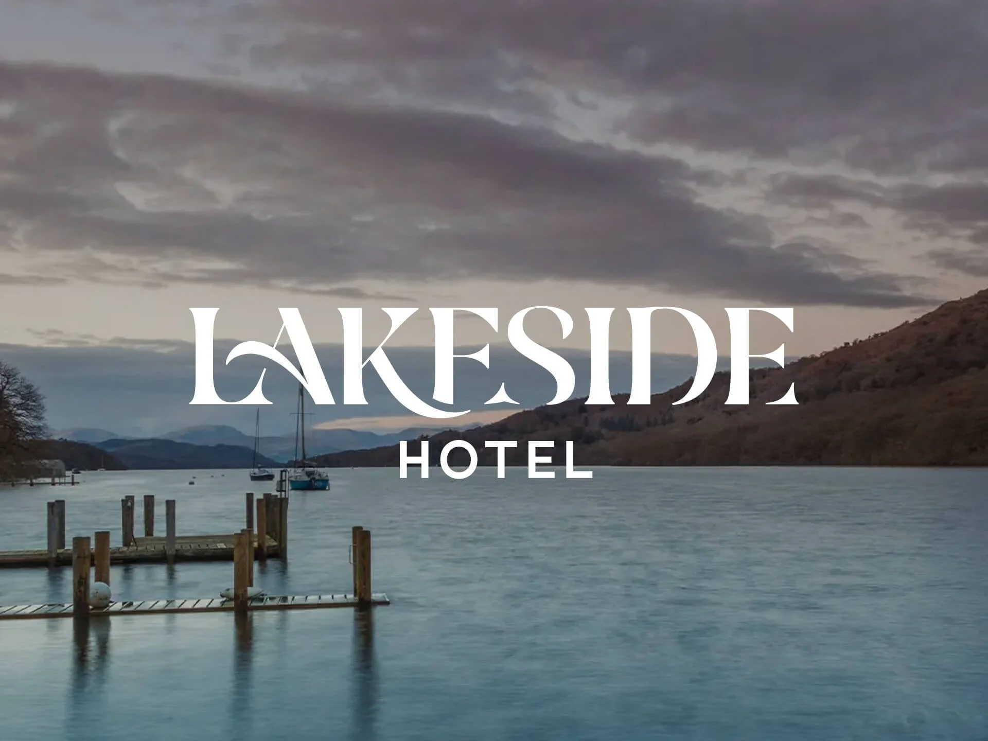 Lakeside Hotel logo over a mountain lake with sailboats and a wooden dock under a cloudy sky.