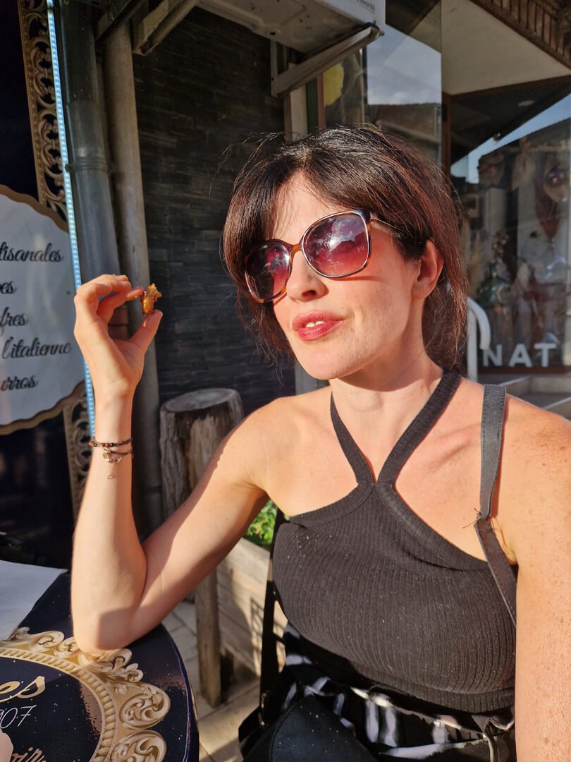 Woman with short brown hair and sunglasses sitting outside at a table, holding a small piece of food, smiling, in bright sunlight.