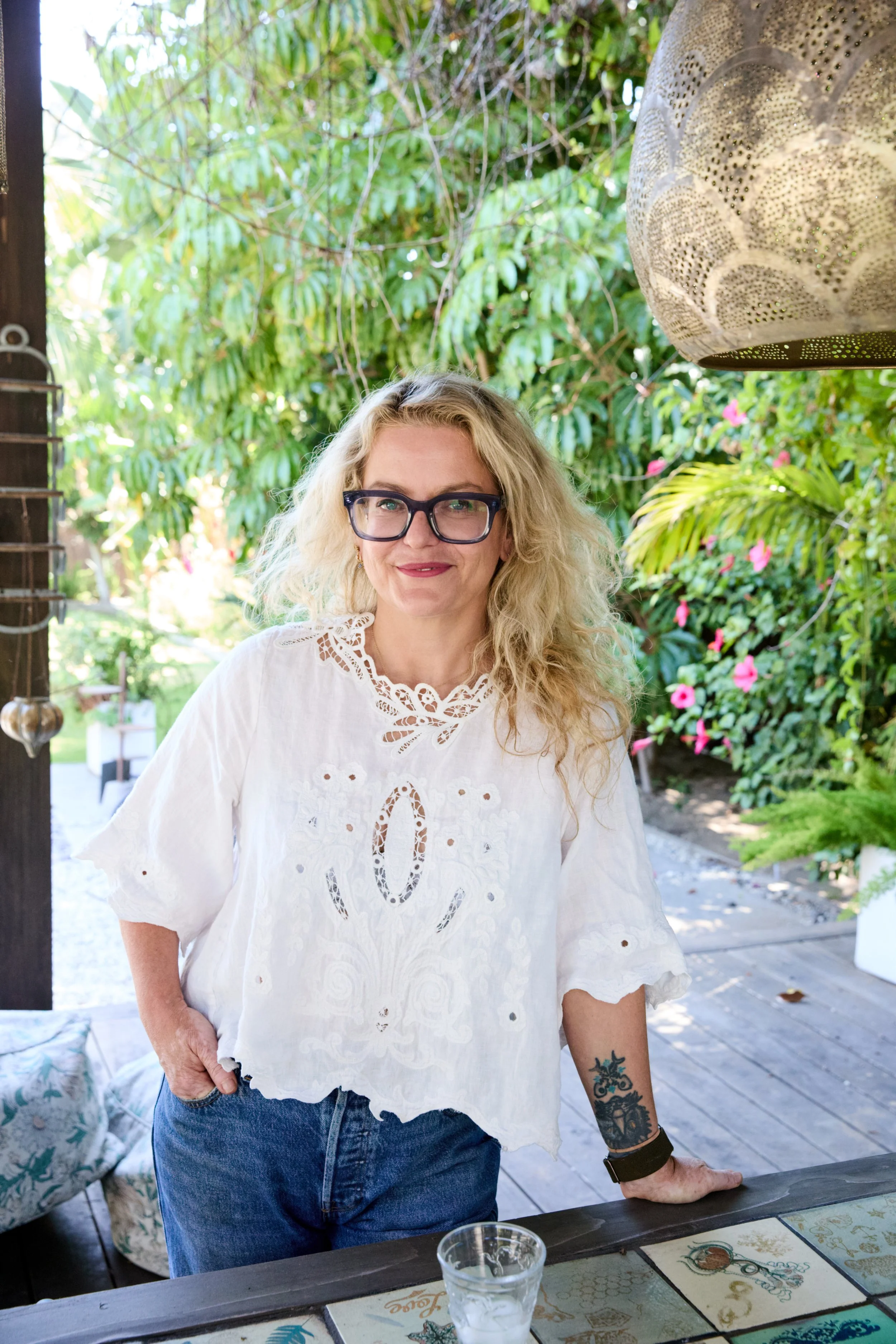 A woman with blonde curly hair, glasses, and tattoos on her arm standing outdoors at a wooden table with colorful placemats, surrounded by lush green foliage and pink flowers.