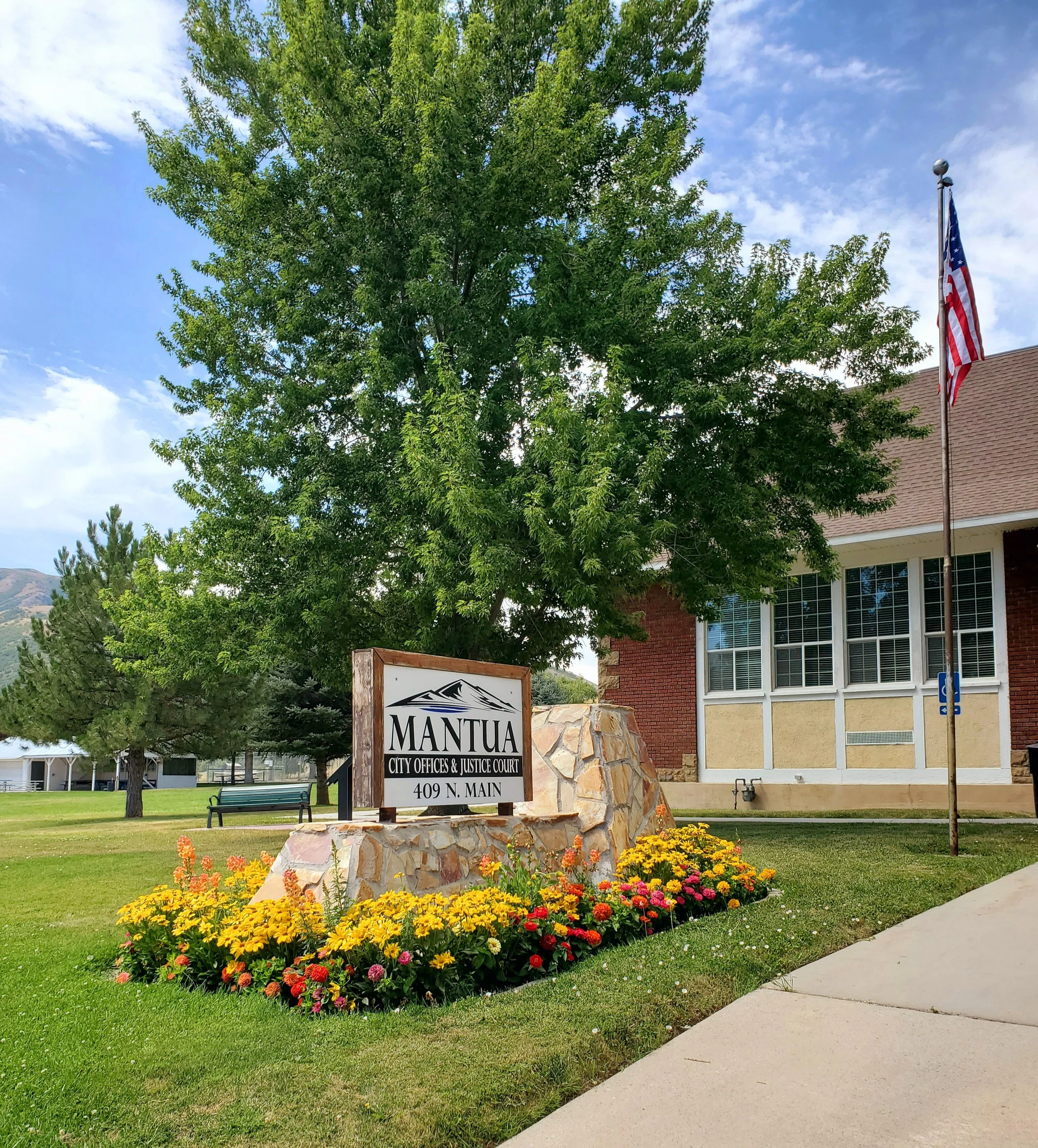 Sign for Mantua City Offices and Justice Court at 409 N. Main, located next to a flower bed and a flagpole with the American flag. A large tree and a building with windows are in the background.