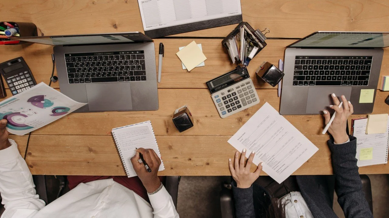 Wooden desk with laptops, papers, other office supplies, and two people working.