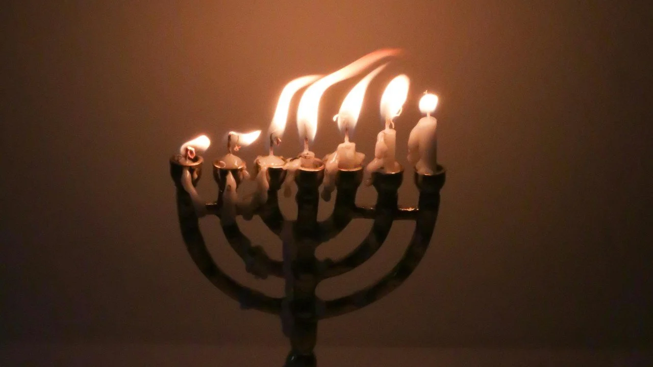 Menorah with seven burning candles, blowing in the wind, with some nearly being put out, against dim, dramatic lighting.