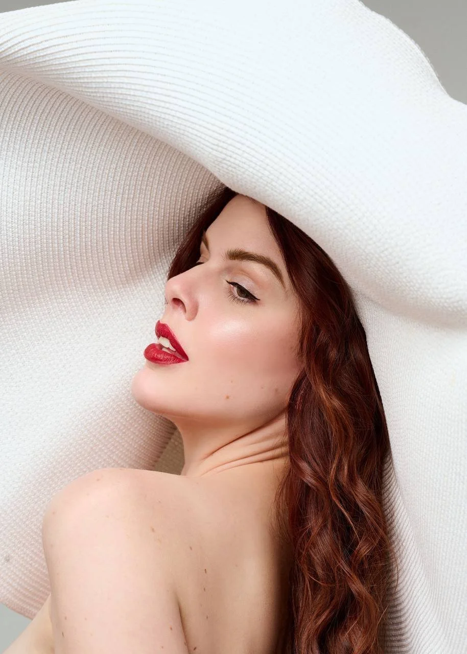 Close-up image of a woman with long, wavy red hair and makeup, wearing a large white hat.