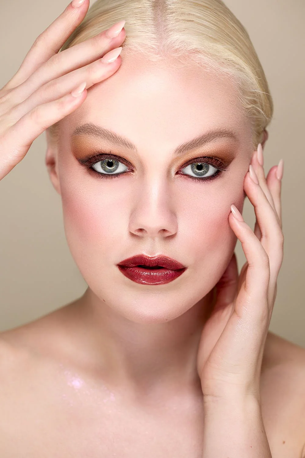 A close-up portrait of a woman with platinum blonde hair and striking makeup, touching her forehead and cheek with both hands.