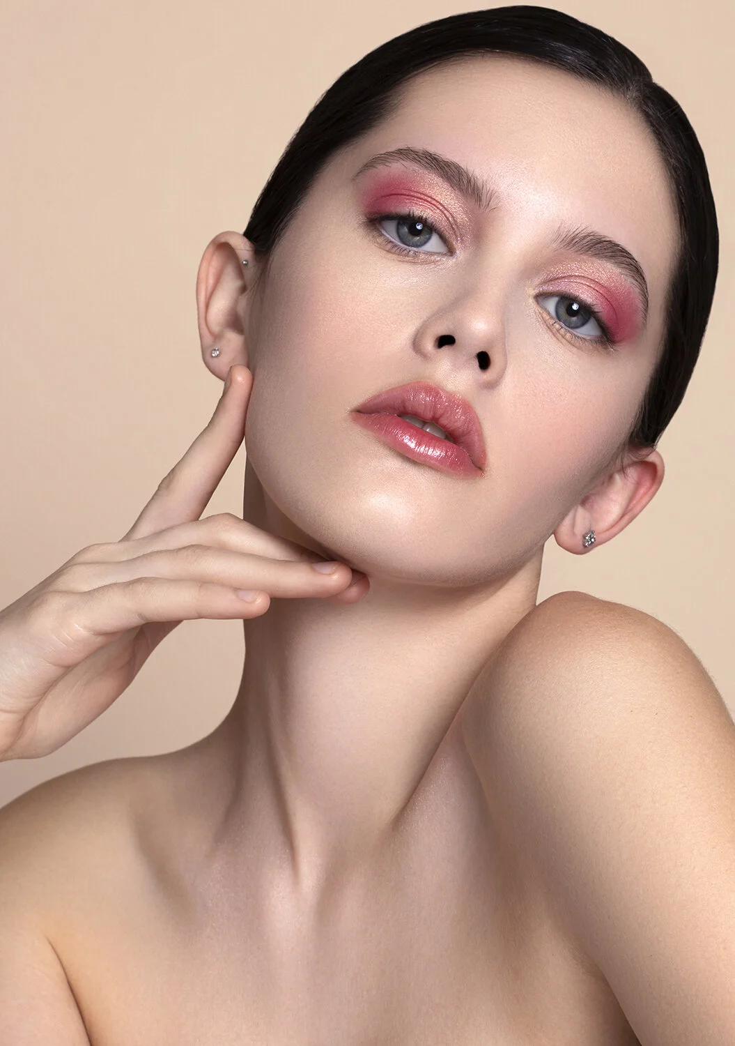 Close-up of a young woman with dark hair, pink makeup, glossy lips, touching her face with one hand, showing clear skin and earrings, against a neutral background.