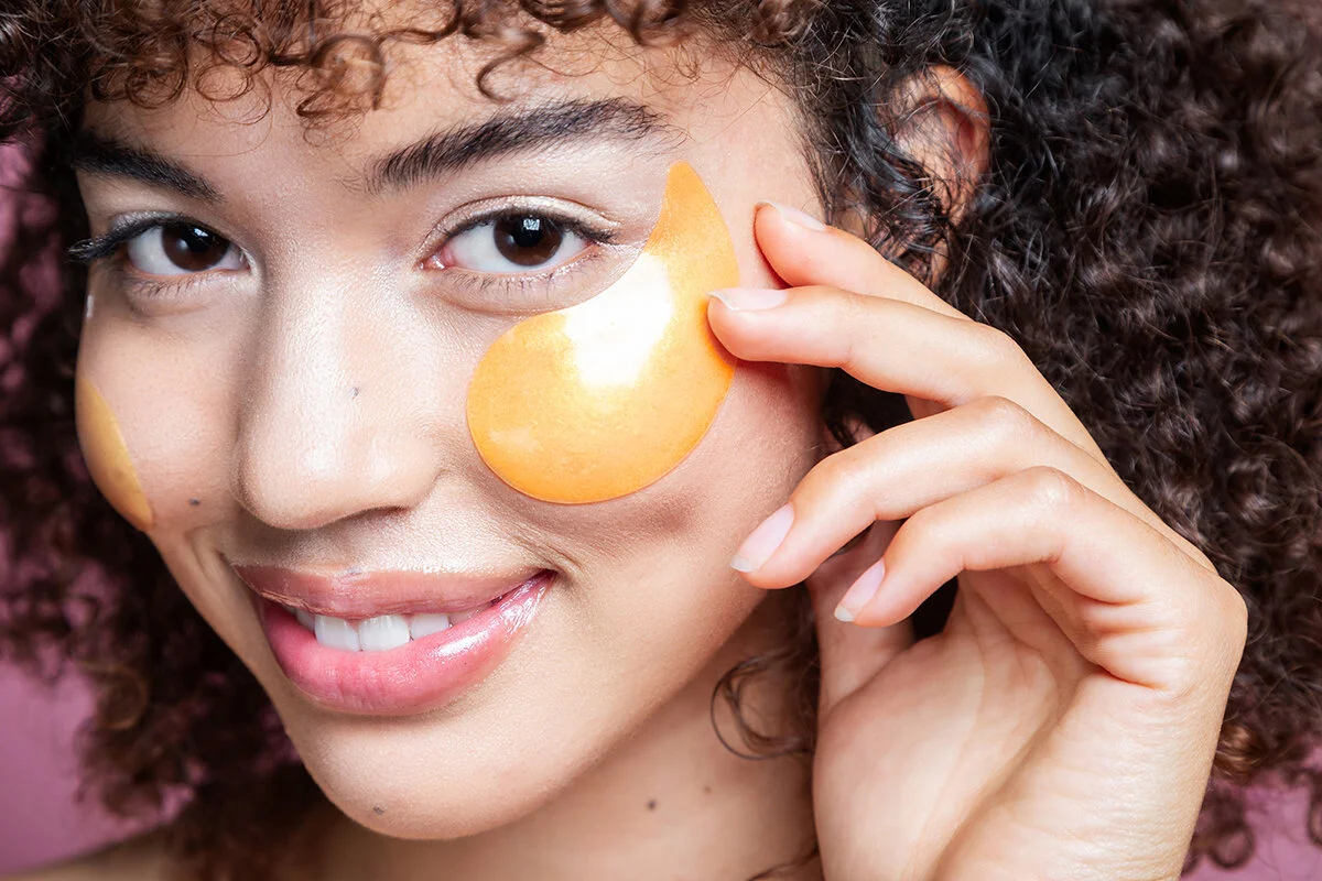 A woman with curly hair holds under-eye patches on her face and smiles.