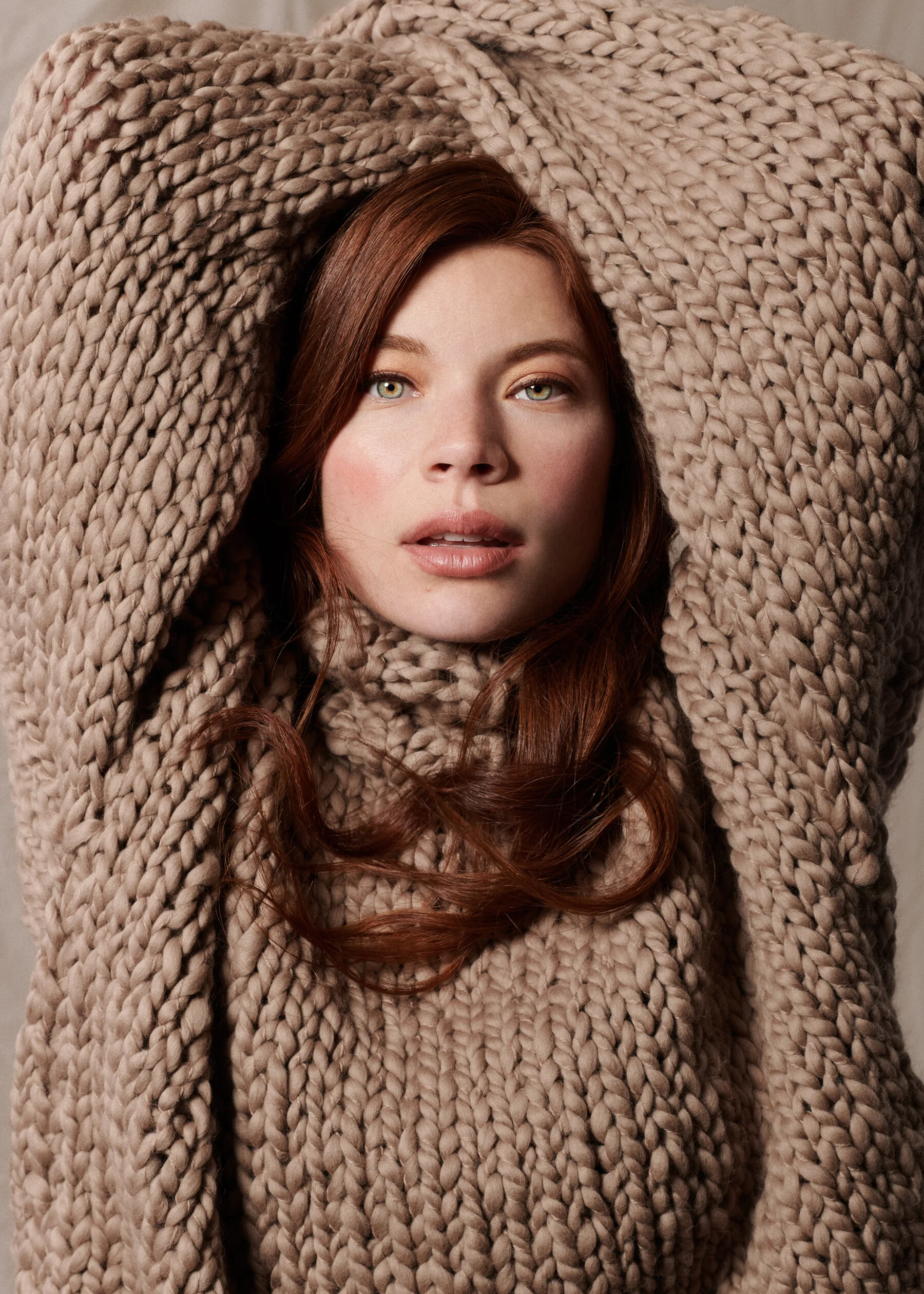 A woman with long red hair wearing a large beige knitted sweater with her arms raised above her head, looking into the camera.