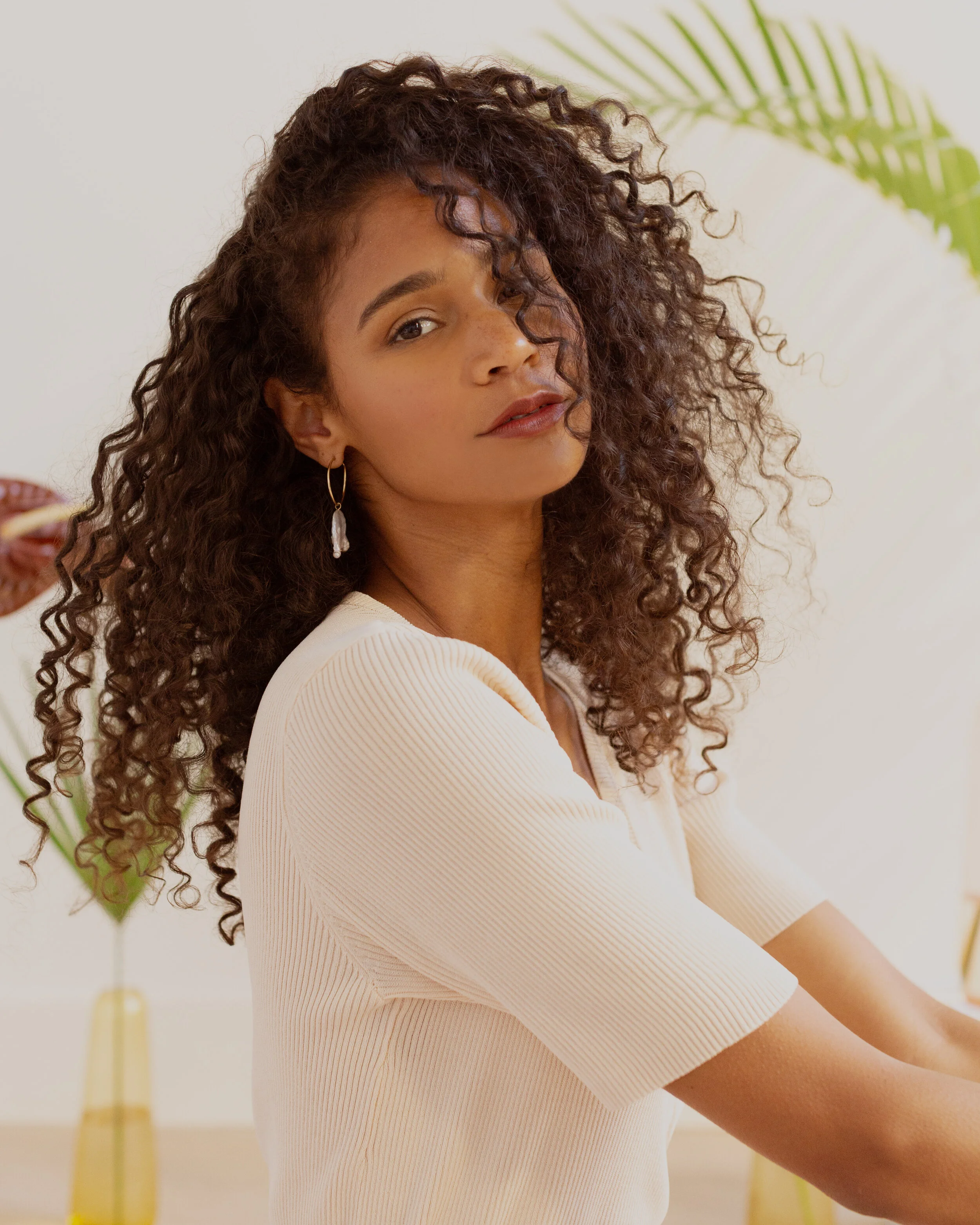 A woman with curly hair wearing a cream-colored top and earrings is looking at the camera. She is indoors with a white wall and green plants in the background.