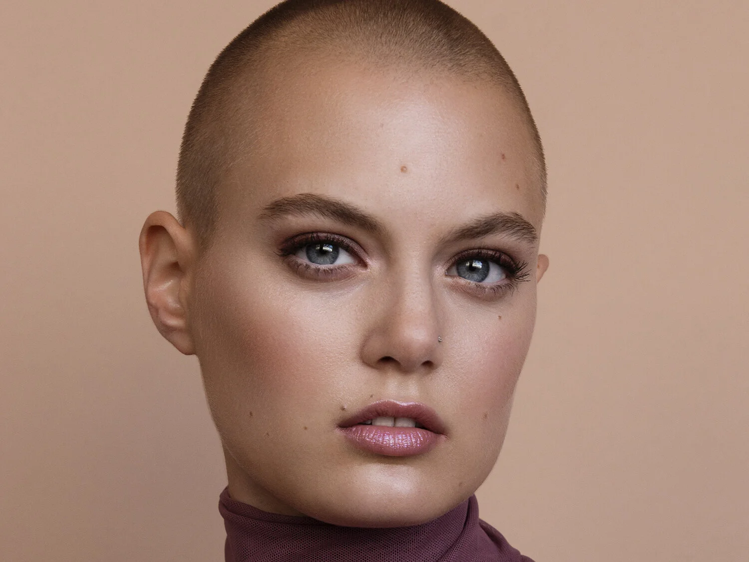 Close-up of a woman with a shaved head, light makeup, and blue eyes, wearing a maroon turtleneck against a beige background.