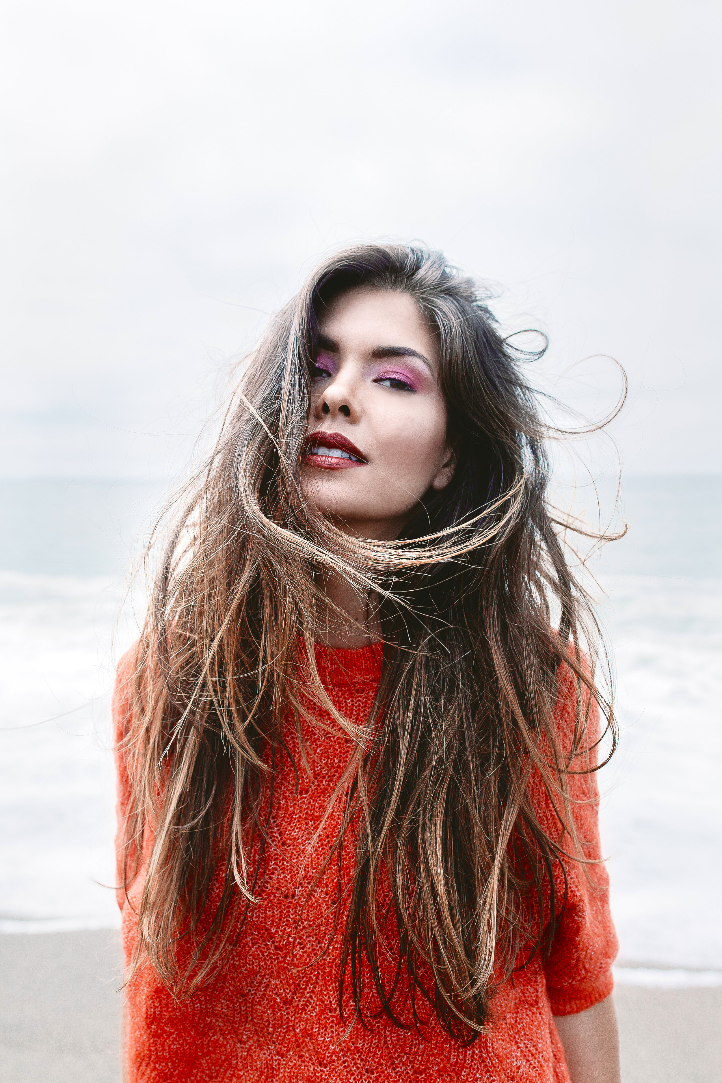 A woman at the beach with windblown hair, wearing a red sweater, looking confidently at the camera.