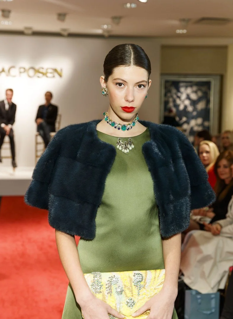 A model on a runway wearing a green dress, a dark fur stole, and jewelry, with an audience and a panel of judges in the background.