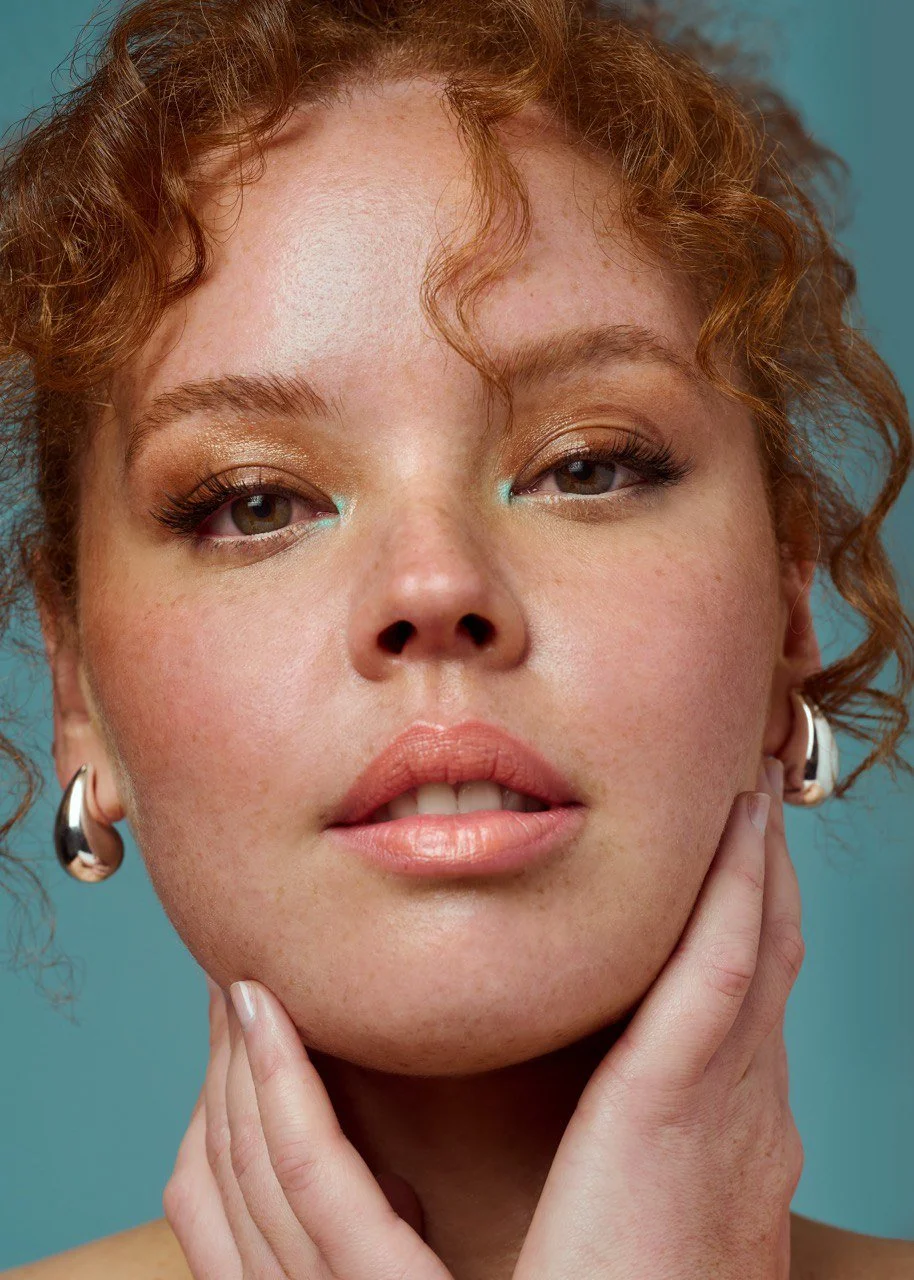 Close-up portrait of a young woman with curly red hair, wearing silver hoop earrings, with makeup featuring gold eyeshadow and nude lipstick, gently touching her chin with her hand against a teal background.