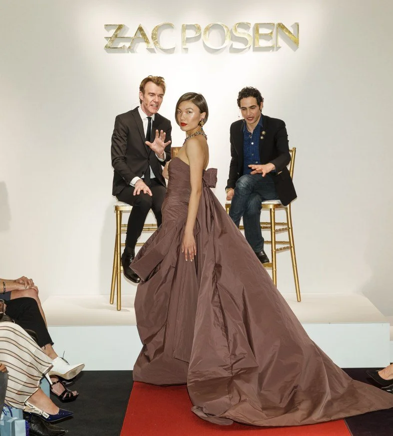 A woman in a strapless, brown gown with a large train posing in front of two men seated on chairs, with a white wall background and the words 'ZAC POSEN' displayed above.