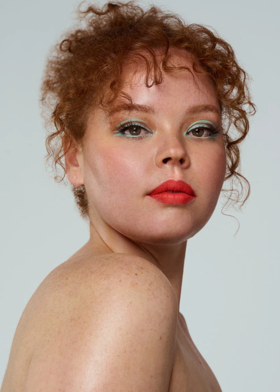 Close-up portrait of a woman with curly red hair, blue eye makeup, and red lipstick, looking at the camera with a neutral expression.