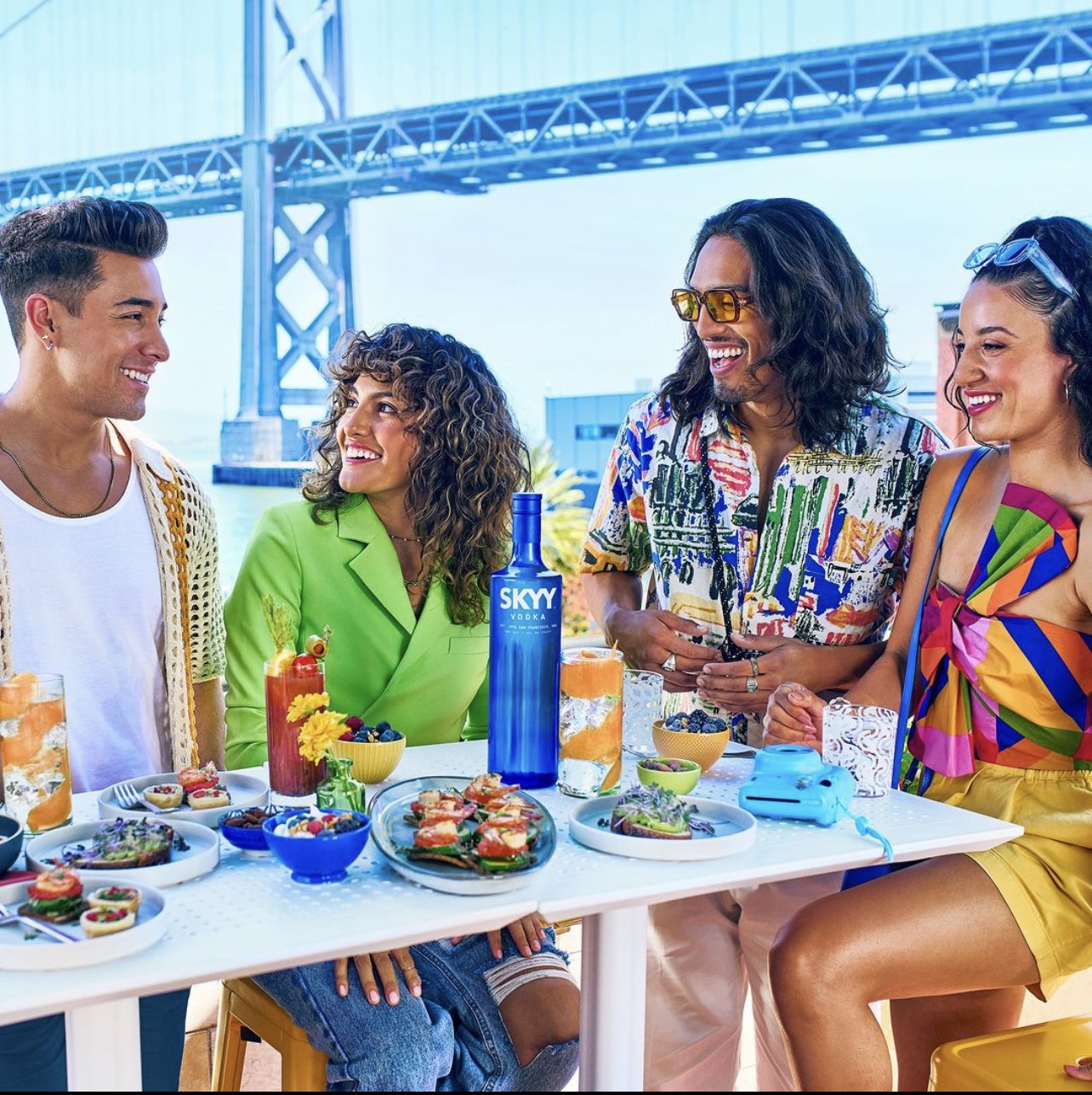 Four friends at a rooftop brunch table with cocktails, snacks, and a bottle of Skyy vodka, under a bridge on a sunny day.