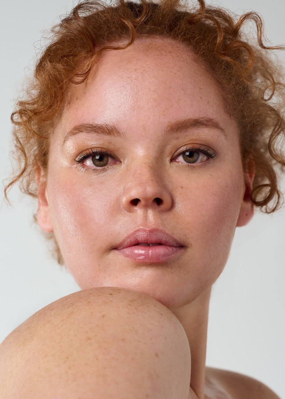 Close-up of a young woman with light skin, freckles, and curly red hair, looking directly at the camera with a neutral expression.