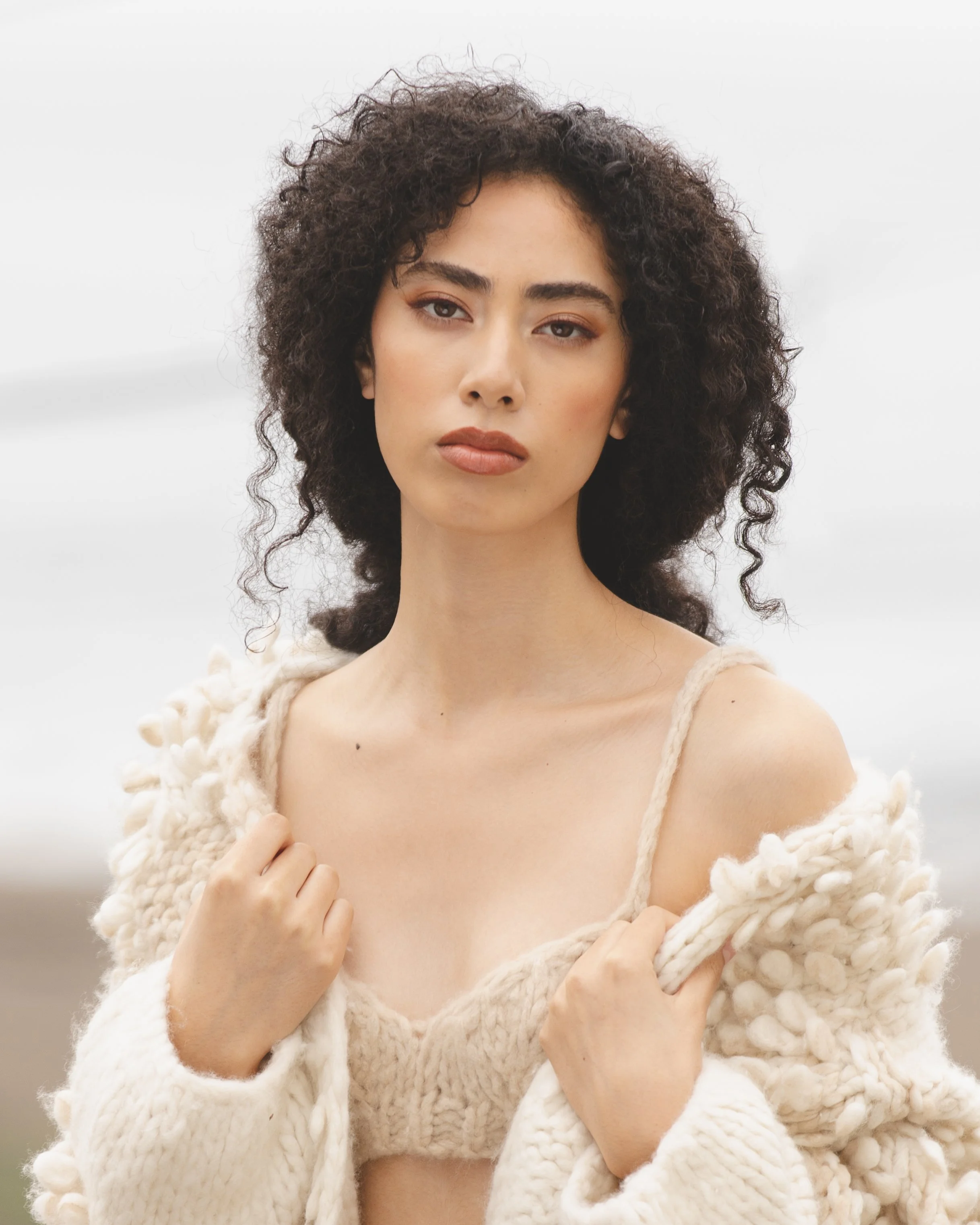 Portrait of a woman with curly dark hair wearing a cream-colored knitted sweater and a matching top, outdoors with a blurred background.