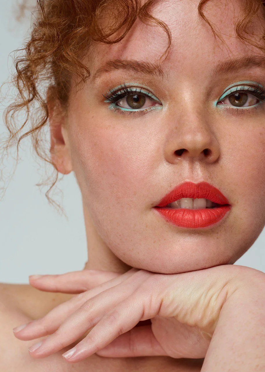Close-up of a woman with red curly hair, makeup featuring blue eyeshadow and red lipstick, resting her chin on her hand.