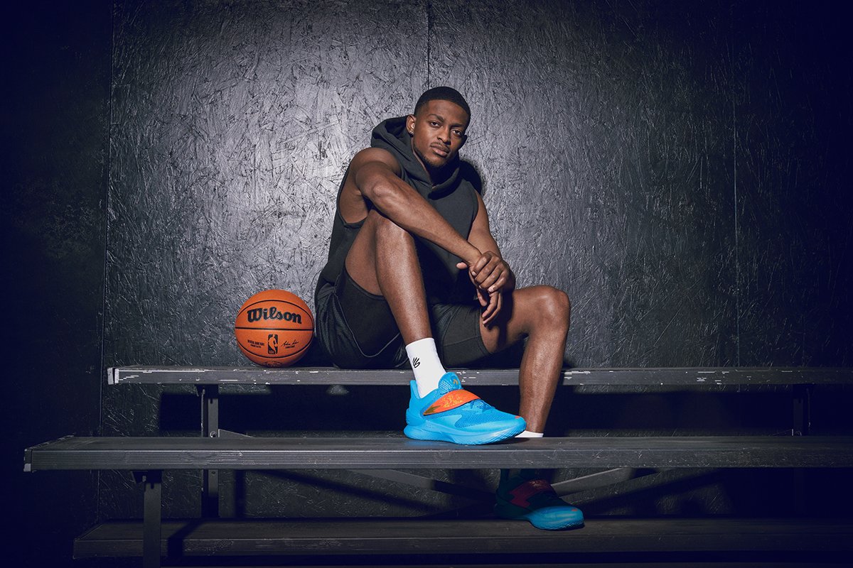 A young man sitting on a bench in a sportswear outfit, with a basketball next to him, in a dark indoor setting.