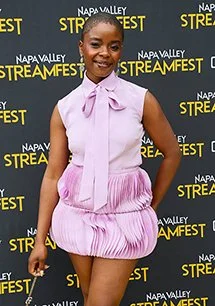 Woman smiling at Napa Valley Streamfest event, wearing a light pink sleeveless dress with a bow tie neckline, standing in front of a black banner backdrop.