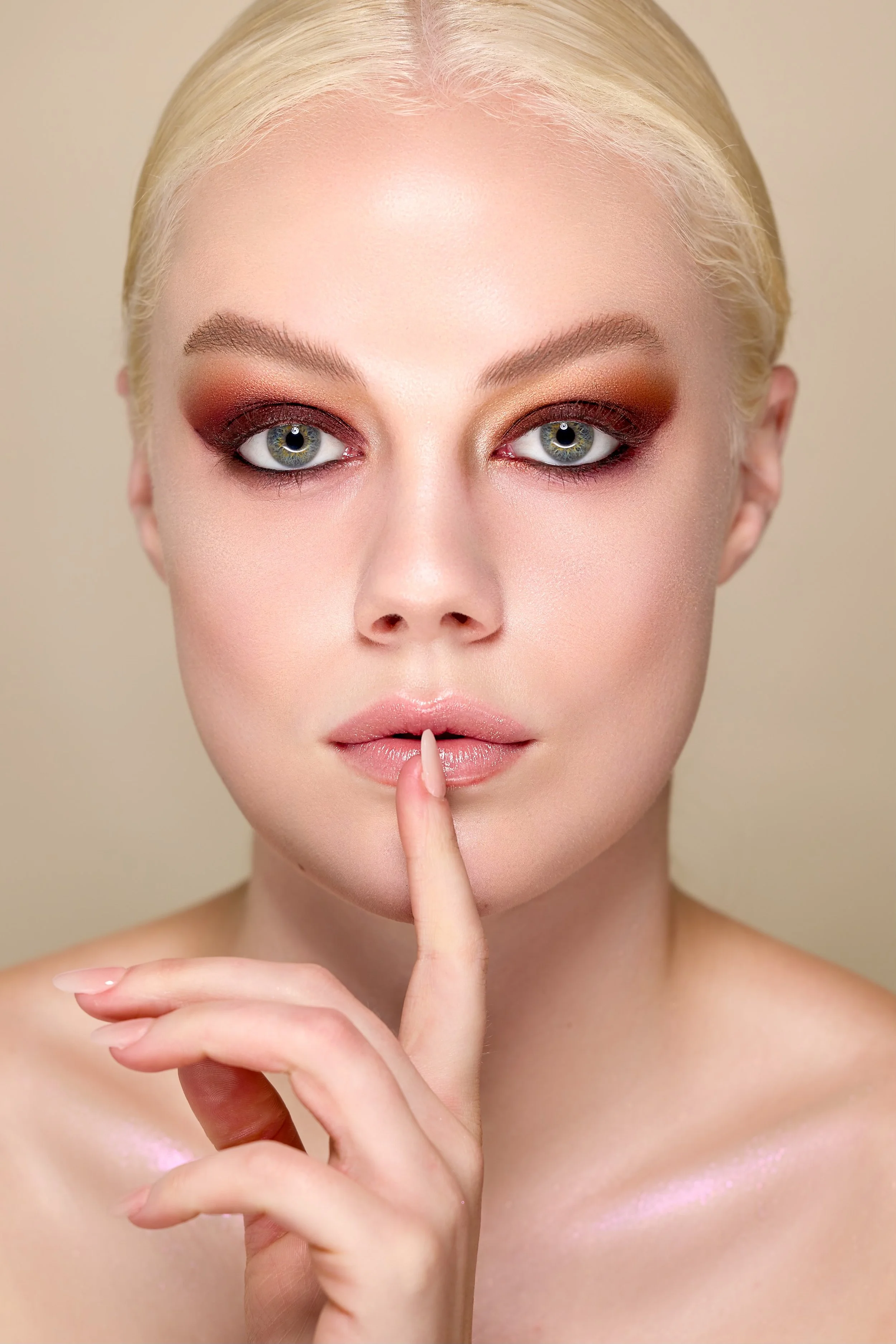 Close-up portrait of a woman with platinum blonde hair, blue eyes, and makeup with warm tones, holding her index finger to her lips in a shushing gesture.
