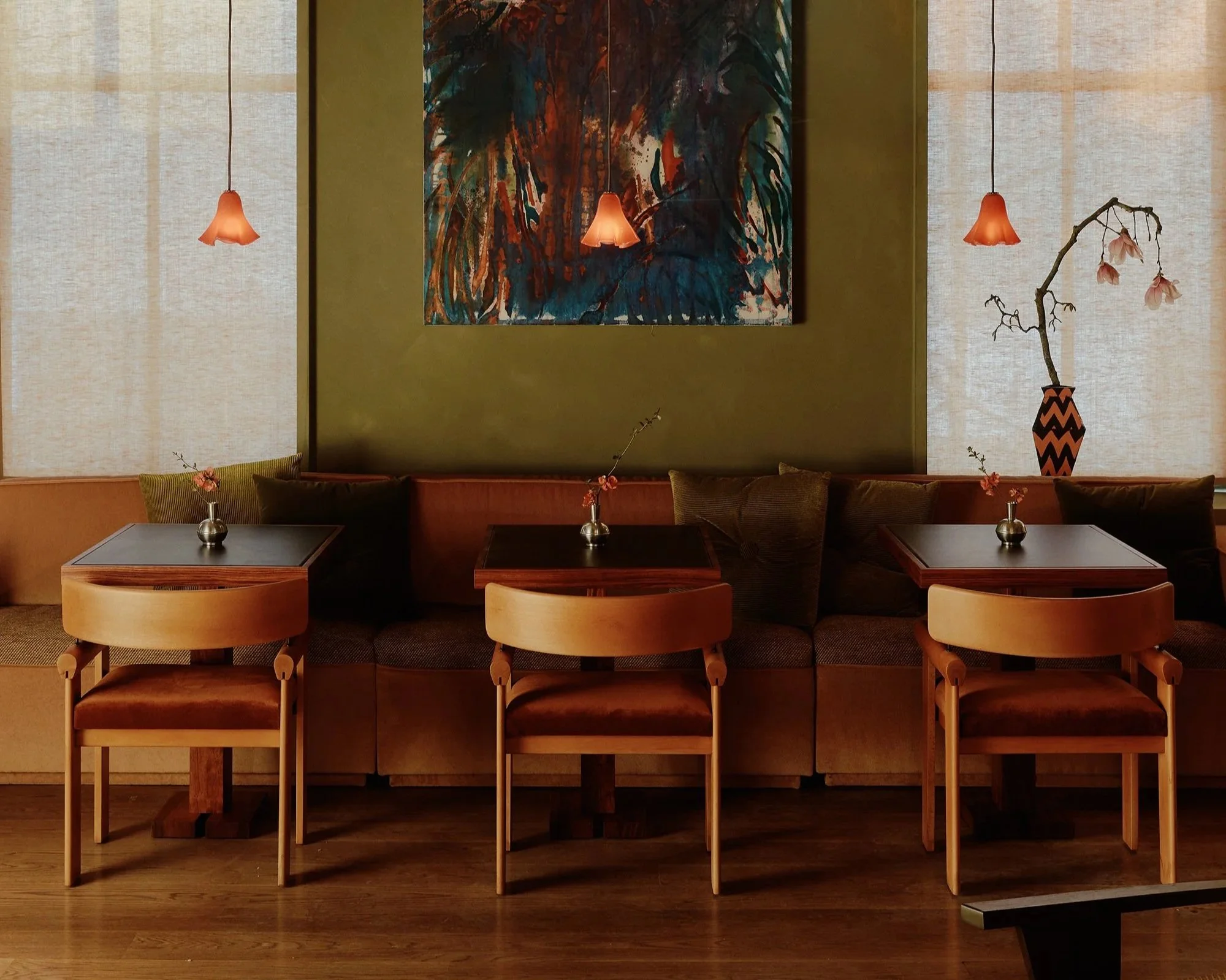 A cozy dining area with three wooden tables, each accompanied by matching chairs, set against a green accent wall. Small vases with delicate flowers sit on each table. Above, three pendant lights with flower-shaped shades hang, illuminating abstract 
