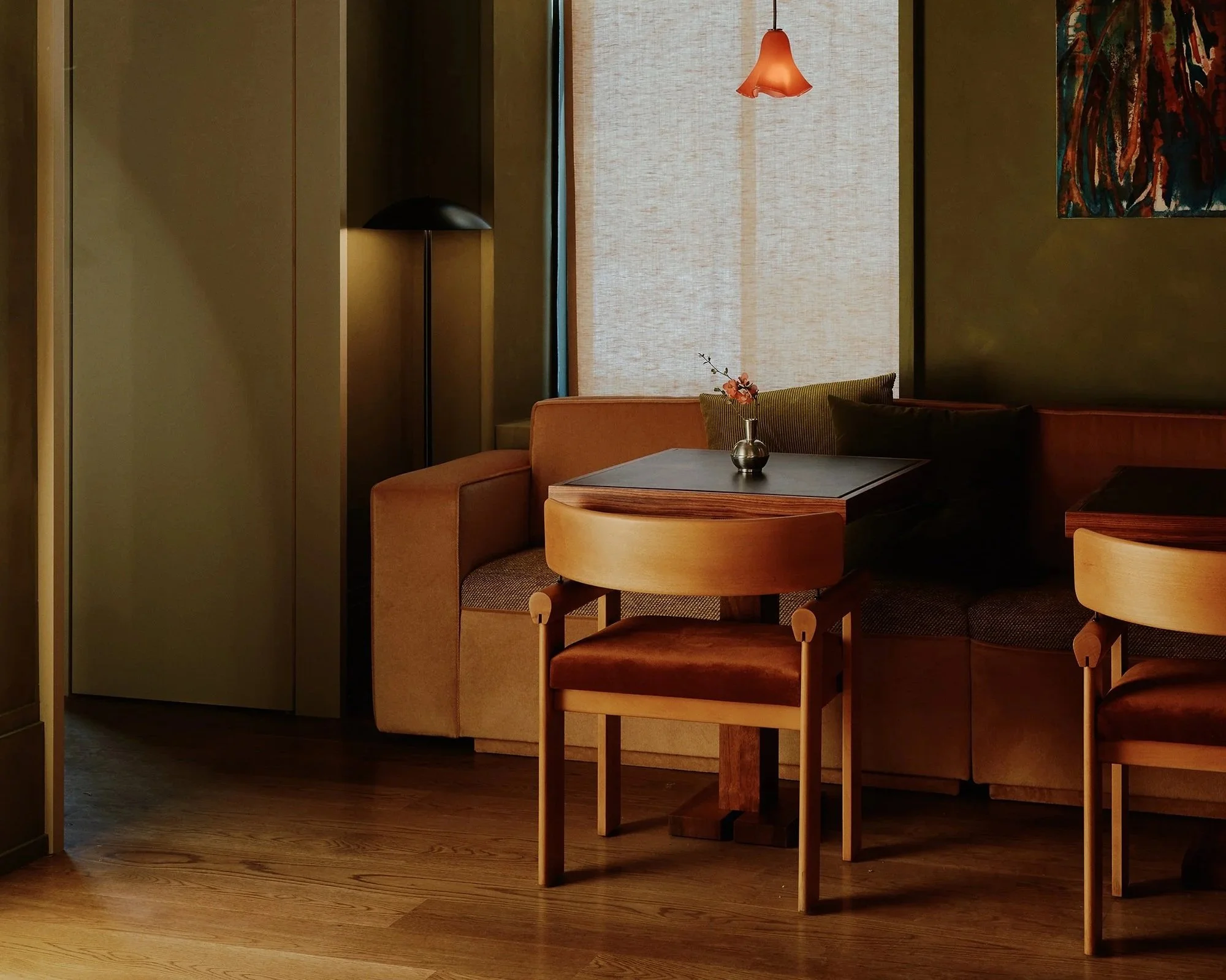 A cozy interior corner with a beige sofa, a wooden dining table, and chairs. A small vase with flowers sits on the table, with warm lighting from a hanging lamp and a floor lamp, and artwork on the wall.