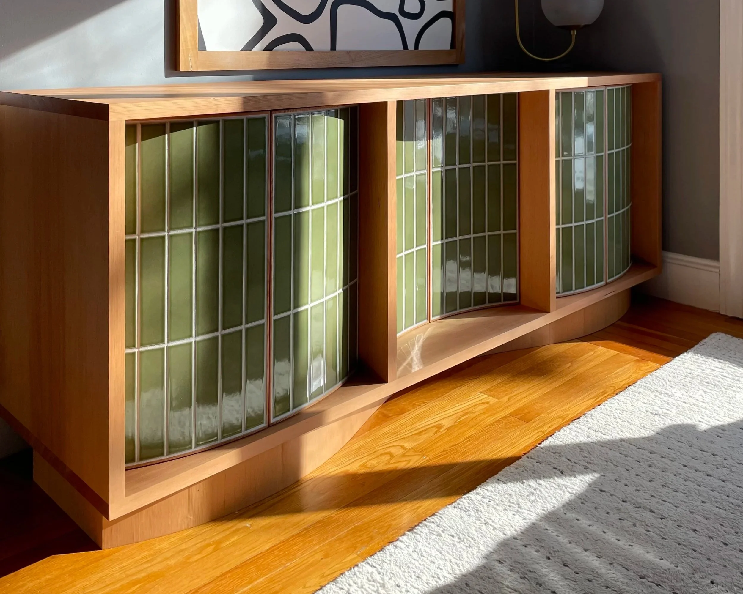 A wooden cabinet with curved ceramic tile panels on the front, placed on a wooden floor next to a white rug, with a black and white abstract art piece on the wall above.  Custom furniture.  Handcrafted by Milk Furniture LLC. 
