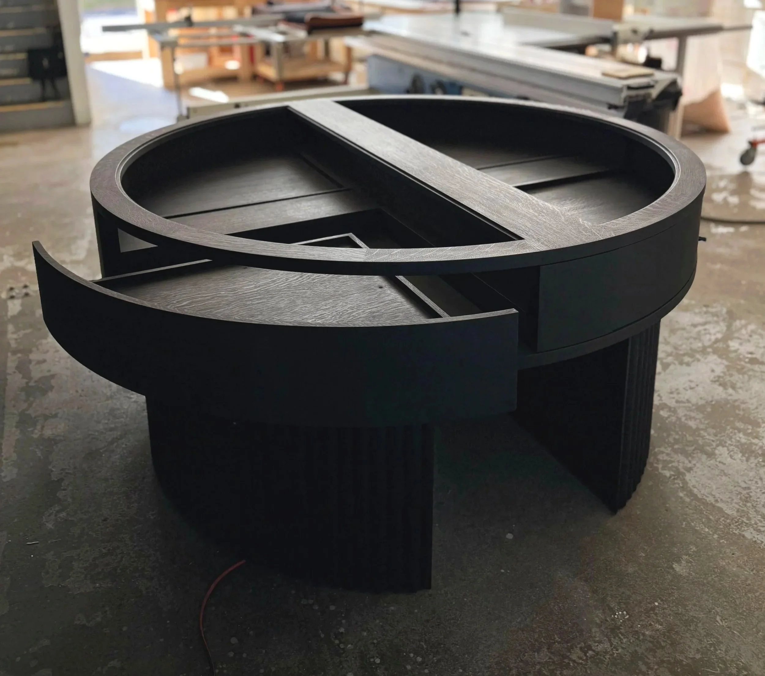 A black circular jewelry case with a unique, modern design featuring an open glass top and curved drawers that open radially. The table is in a workshop with various woodworking tools and materials in the background. Custom Millwork Furniture.