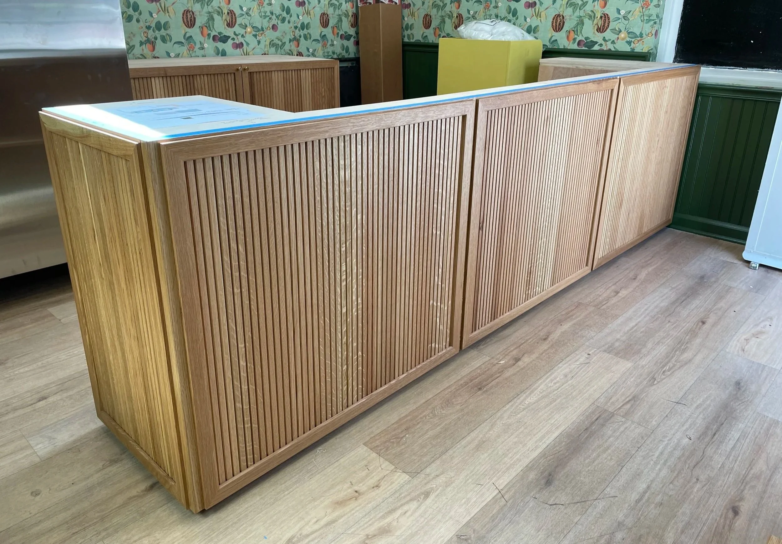 A wooden counter or bar with vertical white oak bevel slats on the front, placed in a room with wood flooring and a colorful wallpapered wall in the background.  Bespoke commercial millwork for Boston Seaport retail.  Custom interior design.