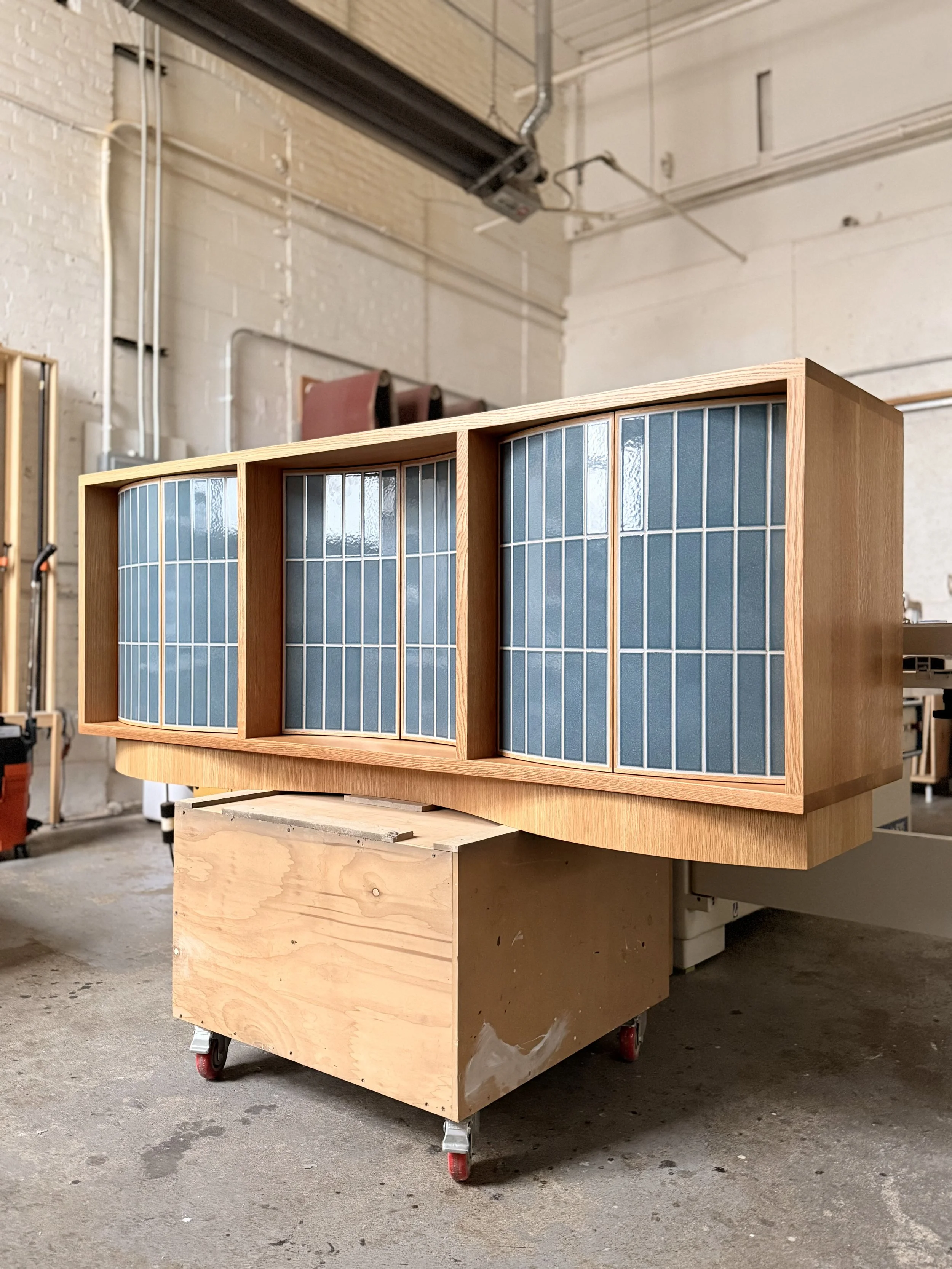 A wooden cabinet with four sections, each with blue glazed ceramic tiles, on wheels in a workshop.  Custom furniture.  Handcrafted by Milk Furniture LLC in New England.