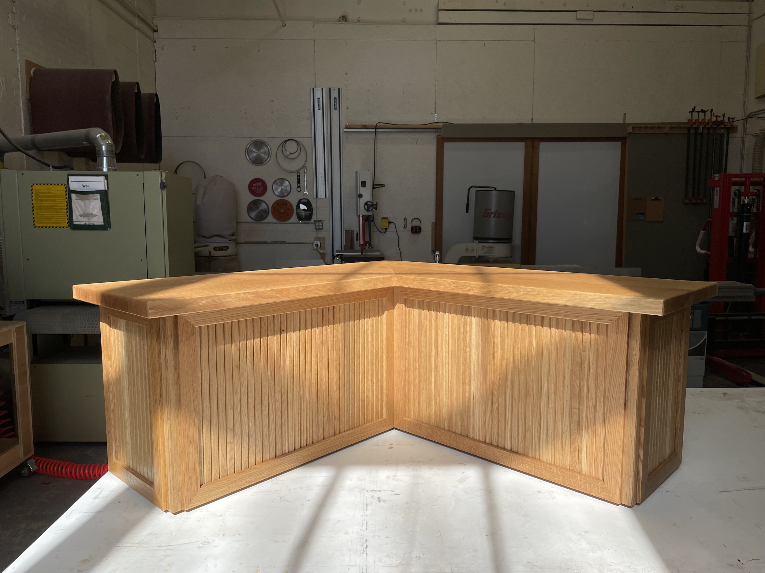 A sun drenched wooden L-shaped bench with vertical wood slats on the sides in framed panels.  White oak construction.  Custom made furniture and millwork handcrafted in New England and Rhode Island.  