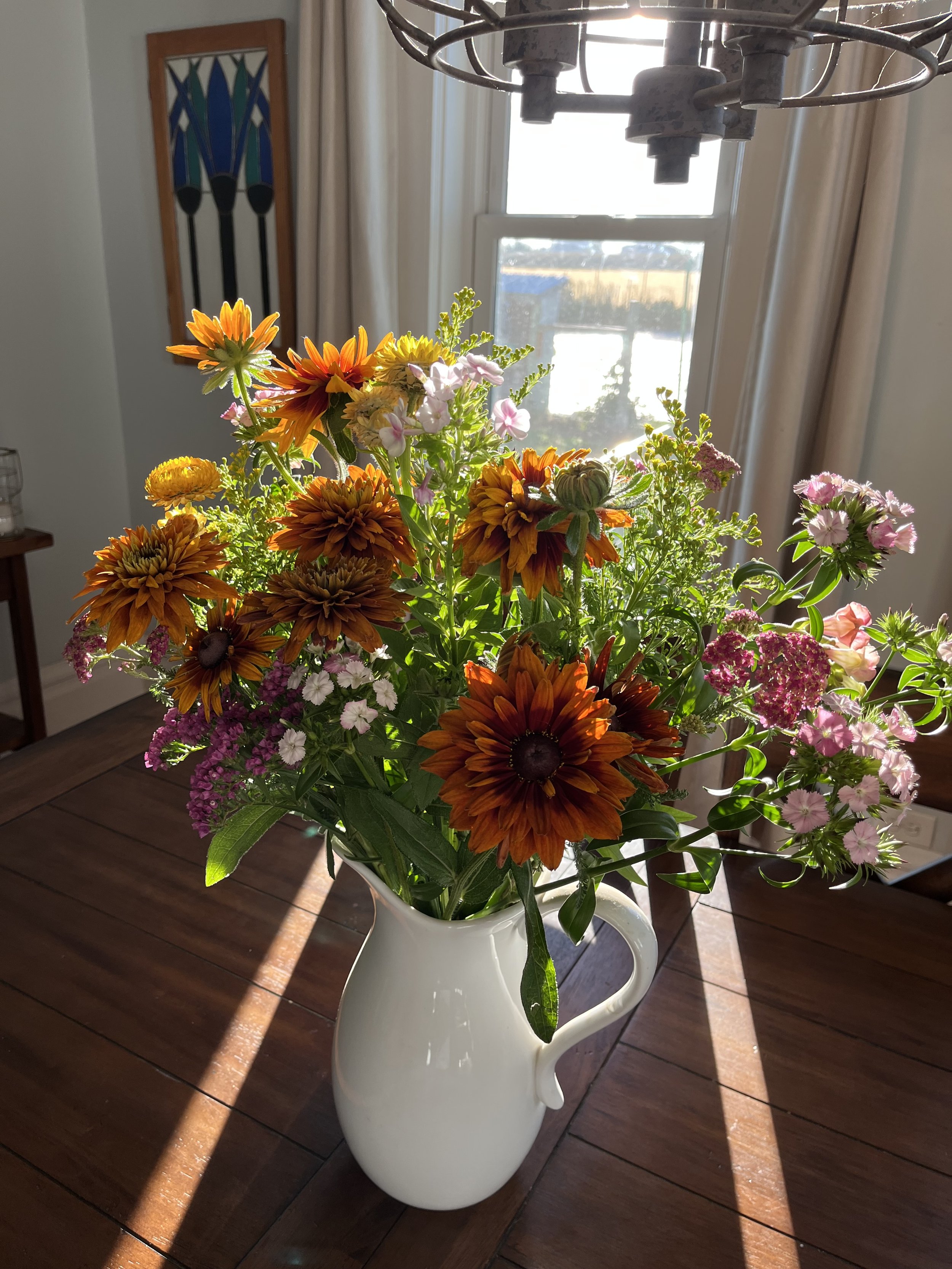 a large bouquet of flowers in a pitcher on a table with the light streaming in