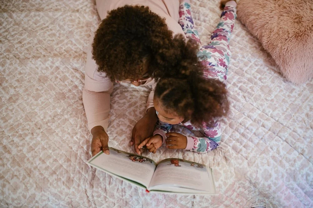 An adult and a child lying on a pink quilted bedspread, looking at a picture book together.