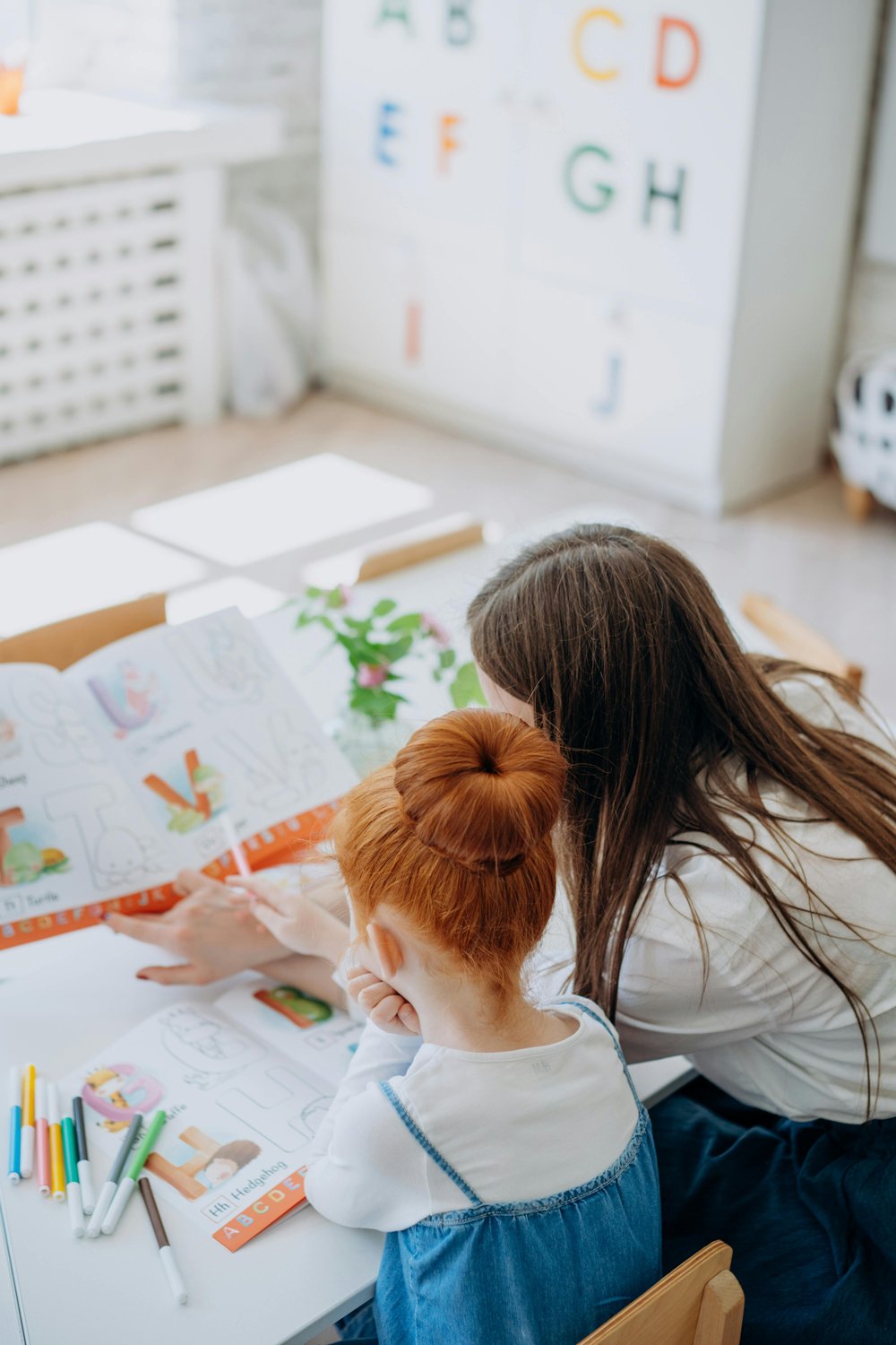 A woman and a young girl with red hair, sitting at a table and looking at a coloring book with colorful illustrations and markers nearby, engaged in a learning activity in a bright room with alphabet posters on the wall.
