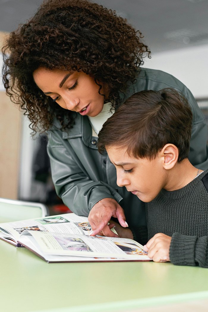 A woman and boy looking at a book together at a table in a classroom.