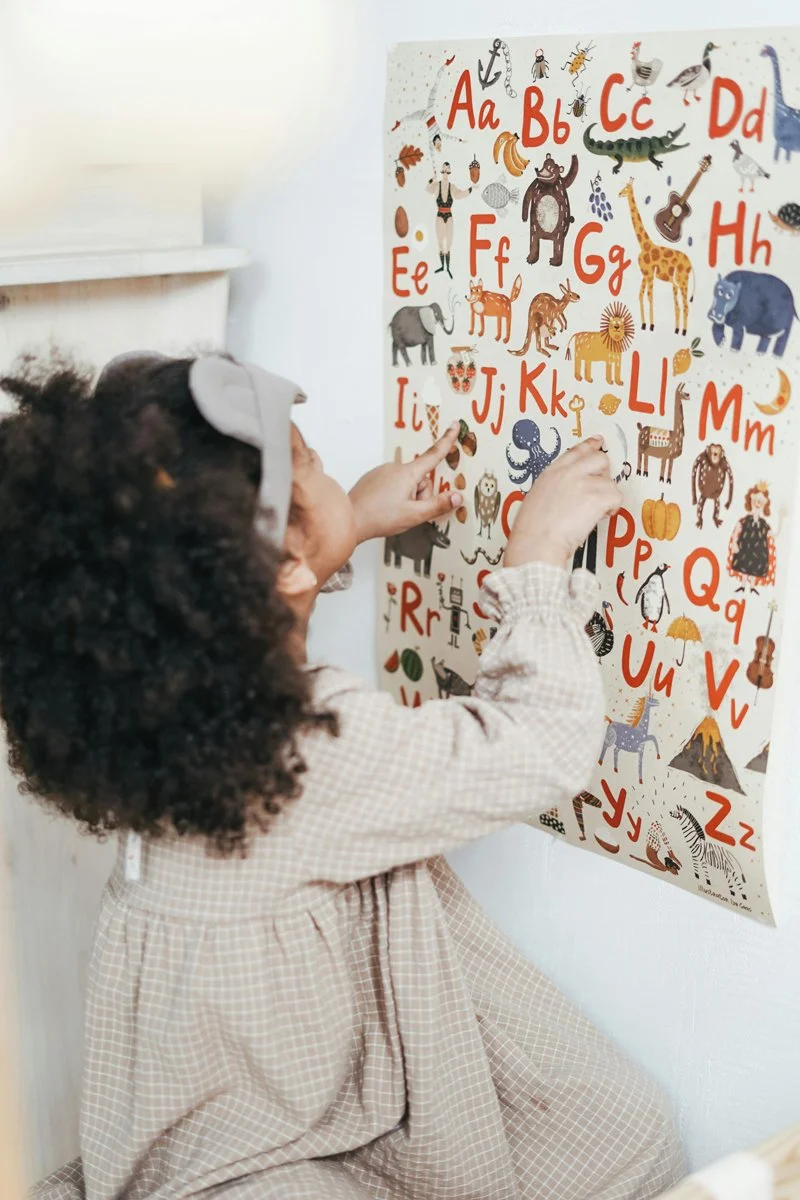 A young girl with curly hair and a bow in her hair is pointing to a colorful alphabet poster on a wall, featuring various animals and objects corresponding to each letter.