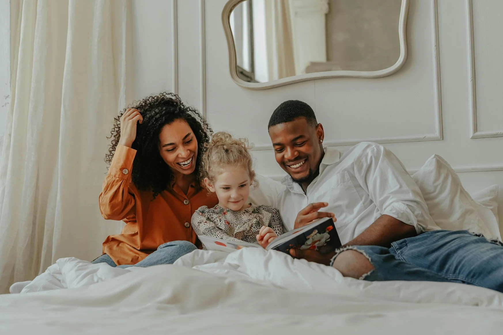 A happy family of three sitting on a bed reading a book together.