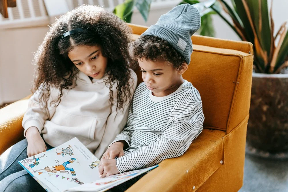 Two children sitting on an orange couch, looking at a book together.