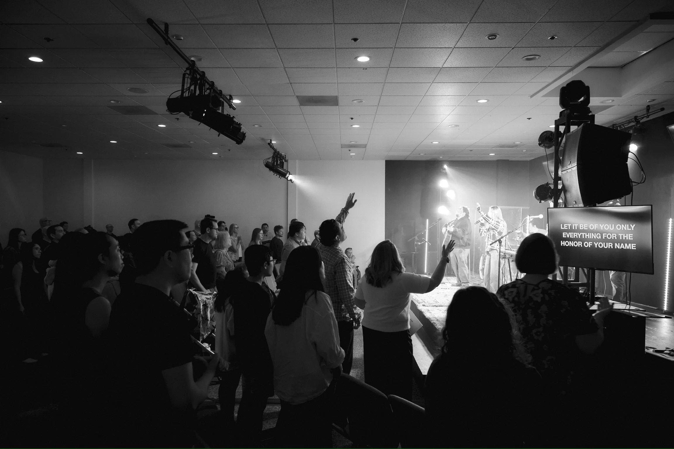 A group of people attending a live music concert, some with hands raised, facing a stage where musicians are performing with bright lights, and a screen displays lyrics.