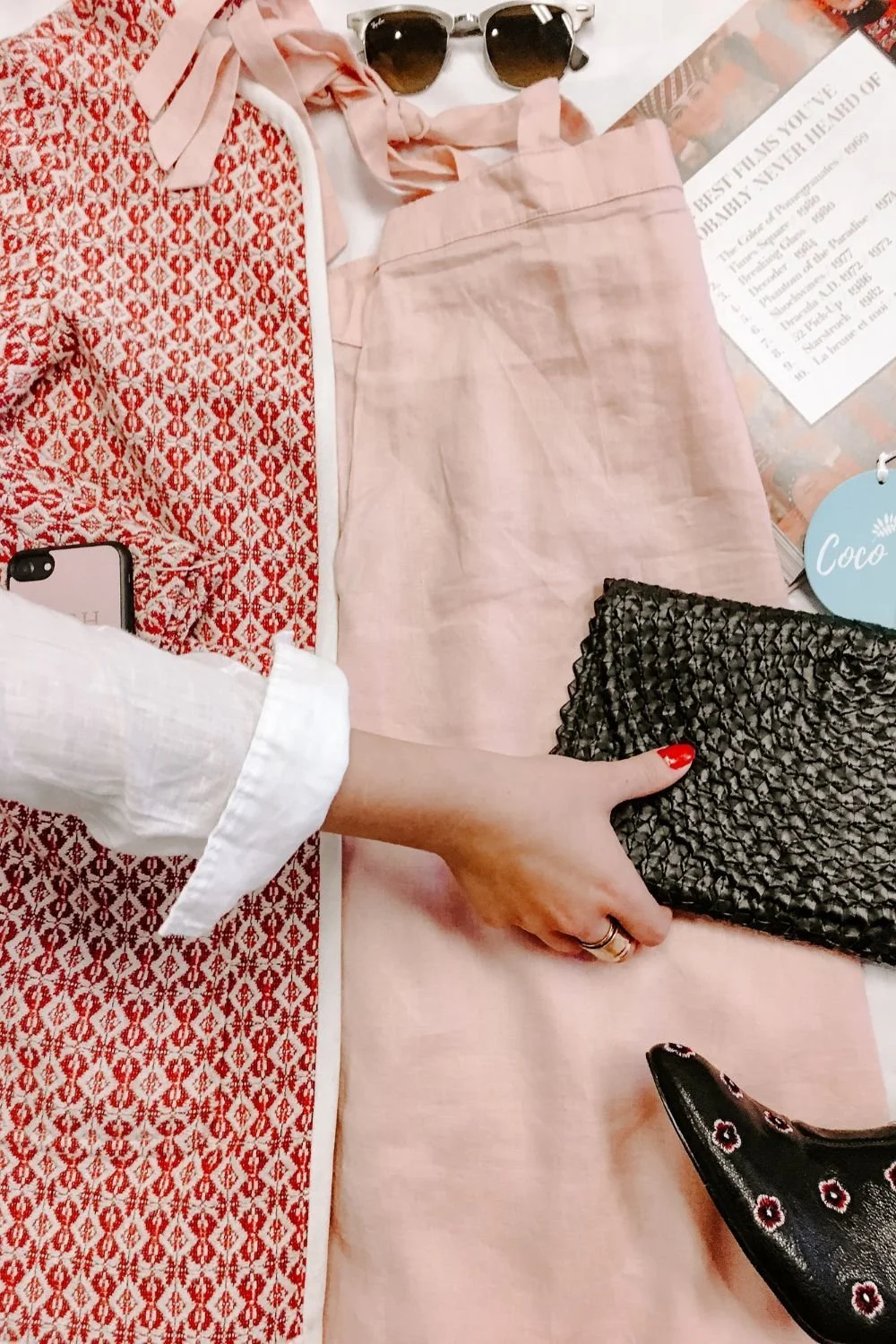 A person with red nail polish is holding a black textured clutch purse. On the table are sunglasses, a pink skirt, a patterned red and white shirt, a magazine, and a black shoe with floral embroidery.