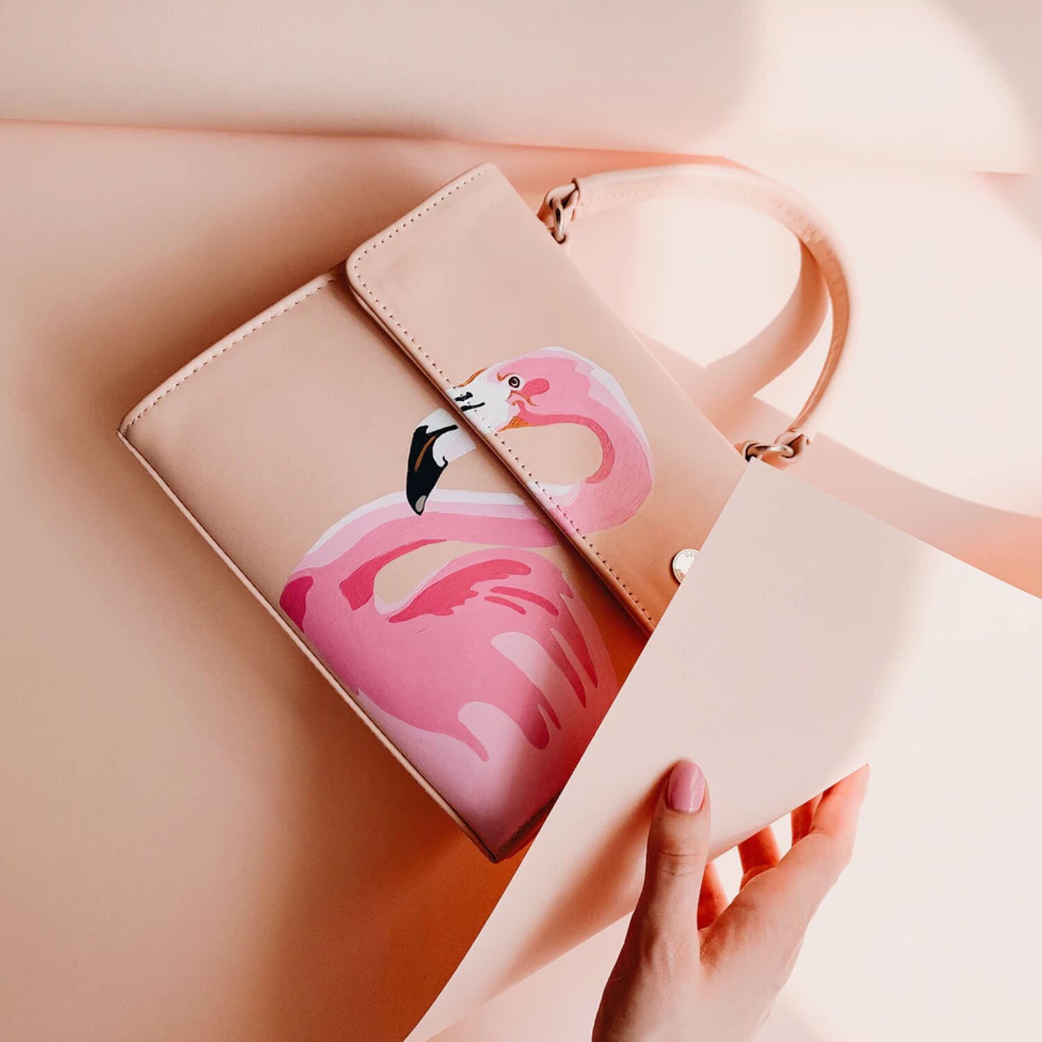 Pink tote bag with a flamingo design on a pink background, partially covered by white paper, held by a person with light skin and pink nail polish.